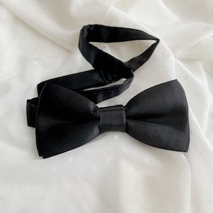 May include: A solid black bow tie with a smooth, satin-like finish. The accessory features a pre-tied bow shape with a central fabric knot and an adjustable black neck strap with a metal slider component for securing the fit around the neck.