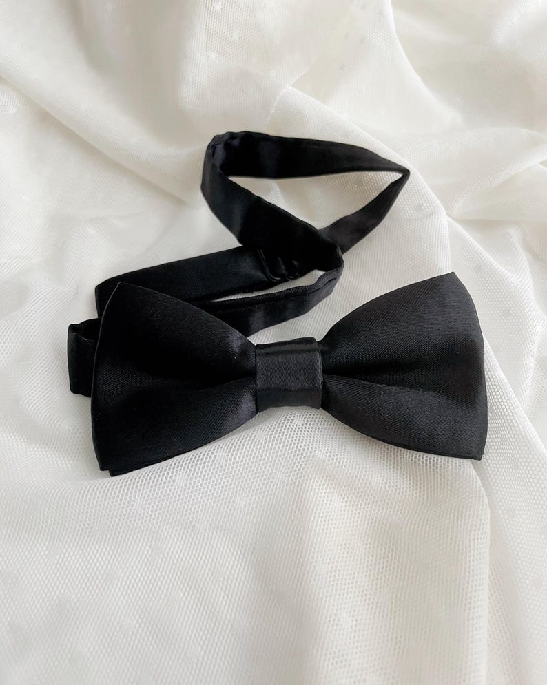 May include: A solid black bow tie with a smooth, satin-like finish. The accessory features a pre-tied bow shape with a central fabric knot and an adjustable black neck strap with a metal slider component for securing the fit around the neck.