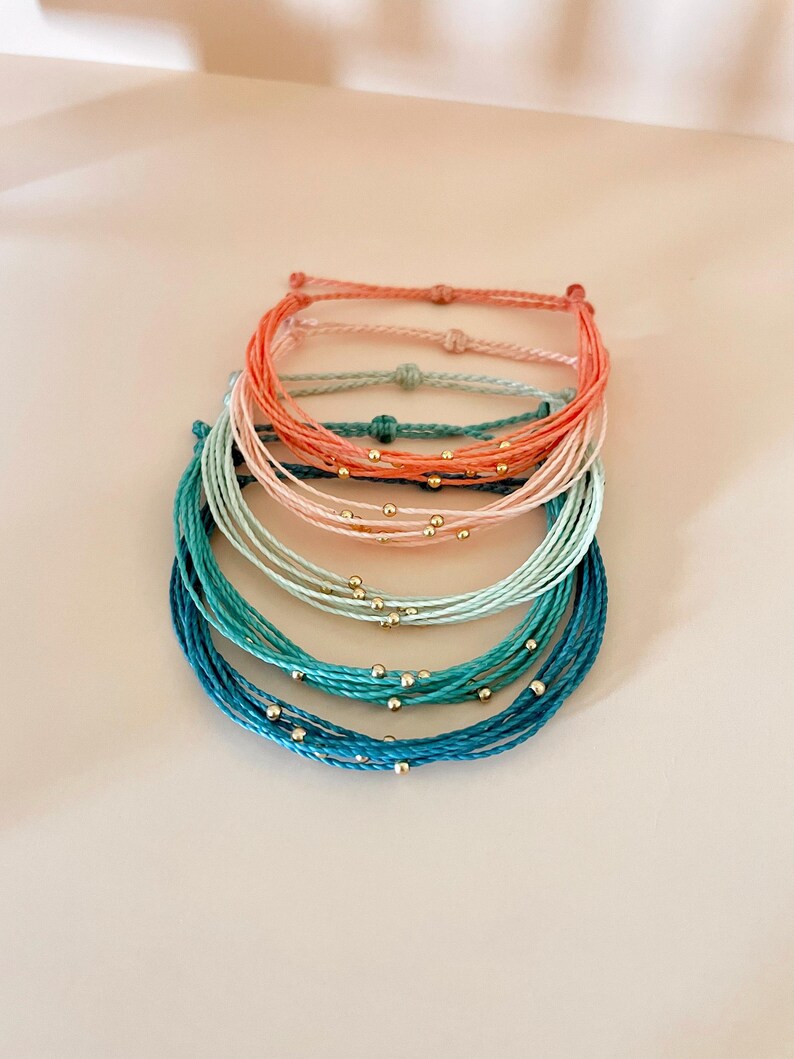K&ouml;nnte beinhalten: Stapel bunter Freundschaftsarmb&auml;nder. Die Armb&auml;nder bestehen aus geflochtenem Kordel in den Farbt&ouml;nen Koralle, Rosa, T&uuml;rkis und Blau. Jedes Armband hat kleine goldfarbene Perlen. Die Armb&auml;nder sind &uuml;bereinander gestapelt.