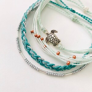 May include: A set of six bracelets featuring a variety of colors and textures, including turquoise, white, and silver. The bracelets are layered and tied together with a silver turtle charm.