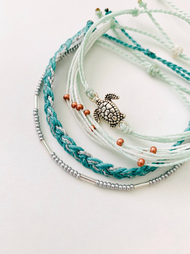 May include: A set of six bracelets featuring a variety of colors and textures, including turquoise, white, and silver. The bracelets are layered and tied together with a silver turtle charm.