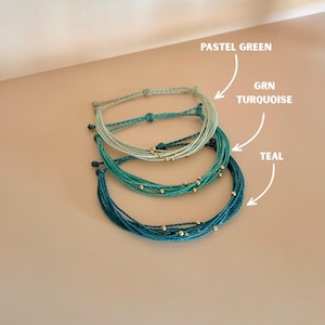 K&ouml;nnte beinhalten: Drei handgefertigte Armb&auml;nder in Pastellgr&uuml;n, T&uuml;rkis und Teal. Jedes Armband besteht aus mehreren Kordelstr&auml;ngen und ist mit kleinen goldenen Perlen verziert. Das Bild enth&auml;lt Etiketten, die die Farben der Armb&auml;nder angeben.