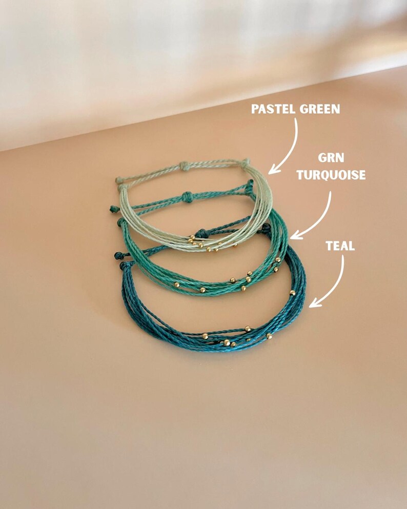 K&ouml;nnte beinhalten: Drei handgefertigte Armb&auml;nder in Pastellgr&uuml;n, T&uuml;rkis und Teal. Jedes Armband besteht aus mehreren Kordelstr&auml;ngen und ist mit kleinen goldenen Perlen verziert. Das Bild enth&auml;lt Etiketten, die die Farben der Armb&auml;nder angeben.