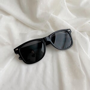 May include: Black sunglasses featuring a classic rectangular frame design with dark, tinted lenses and silver-toned metallic accents on the outer corners of the frame front. The eyewear has a smooth, glossy finish and a standard bridge construction.