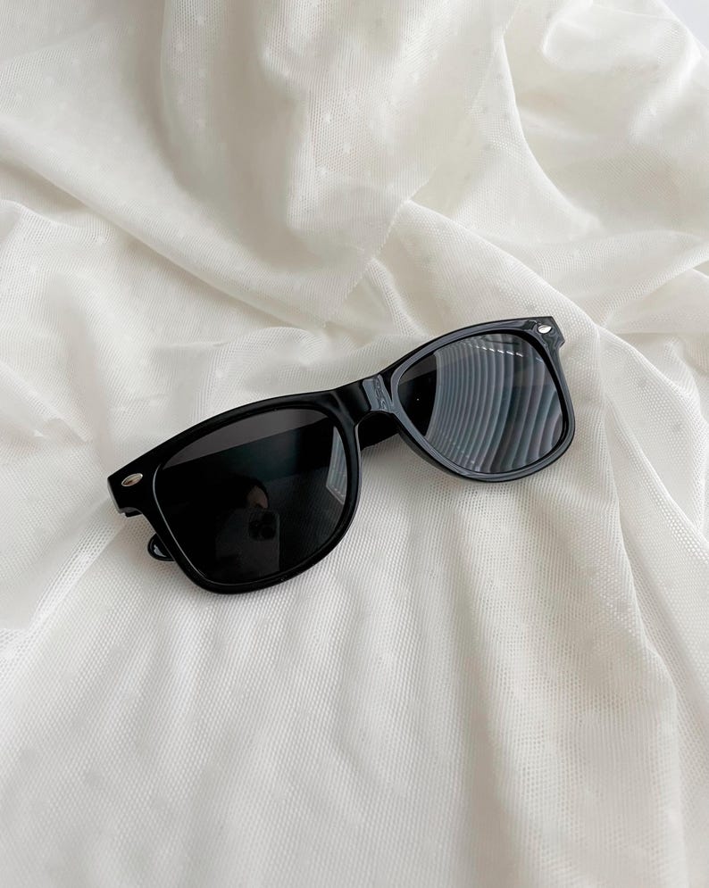 May include: Black sunglasses featuring a classic rectangular frame design with dark, tinted lenses and silver-toned metallic accents on the outer corners of the frame front. The eyewear has a smooth, glossy finish and a standard bridge construction.