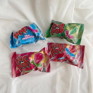 May include: Four individually wrapped Ring Pop candies in blue raspberry, green watermelon, pink strawberry, and red cherry flavors. Each rectangular plastic package features the Ring Pop logo, flavor text, and a graphic of the ring-shaped candy.