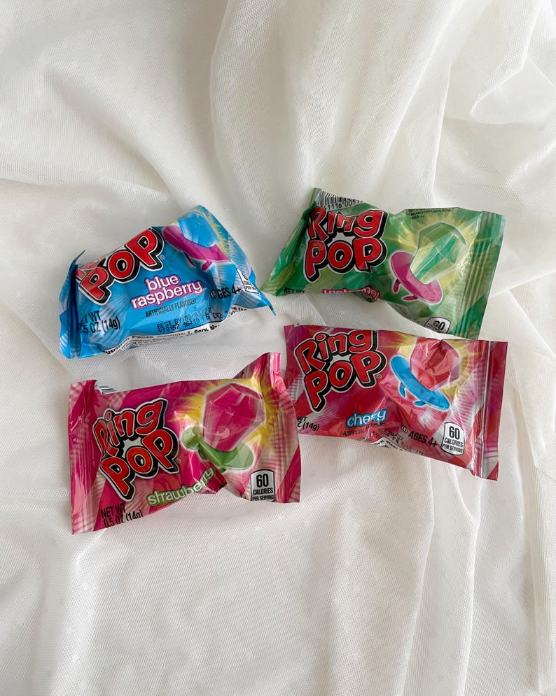 May include: Four individually wrapped Ring Pop candies in blue raspberry, green watermelon, pink strawberry, and red cherry flavors. Each rectangular plastic package features the Ring Pop logo, flavor text, and a graphic of the ring-shaped candy.