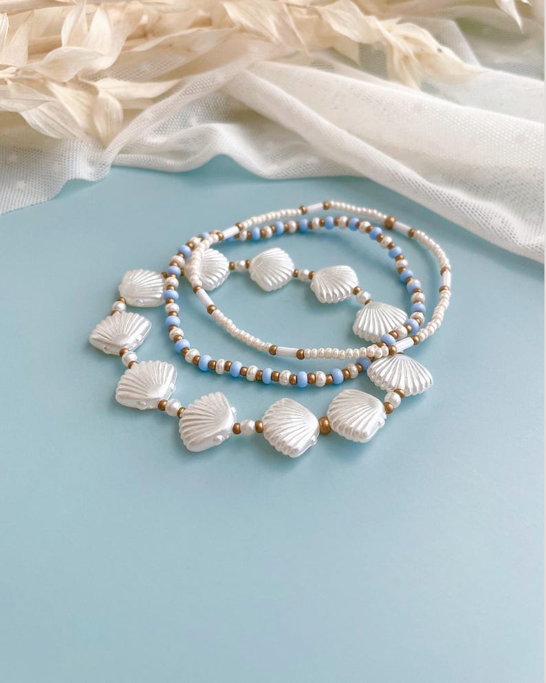 Peut inclure: Un ensemble de trois bracelets de perles compos&eacute; de perles blanches en forme de coquillage, de petites perles rondes bleu clair et de perles intercalaires m&eacute;talliques dor&eacute;es. Les bracelets sont dispos&eacute;s en cercle, montrant un m&eacute;lange de textures.