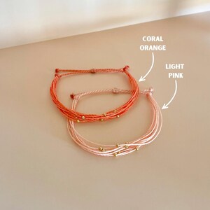 K&ouml;nnte beinhalten: Zwei handgefertigte Armb&auml;nder, eines in Korallenorange, das andere in Hellrosa. Beide Armb&auml;nder bestehen aus mehreren Kordelstr&auml;ngen und haben kleine goldene Perlen. Der Text auf dem Bild lautet "CORAL ORANGE" und "LIGHT PINK".