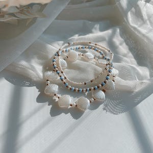 Peut inclure: Un ensemble de trois bracelets de perles compos&eacute; de perles blanches en forme de coquillage, de petites perles rondes bleu clair et de minuscules intercalaires dor&eacute;s m&eacute;talliques. Les bracelets sont dispos&eacute;s en cercle, r&eacute;v&eacute;lant des textures vari&eacute;es.