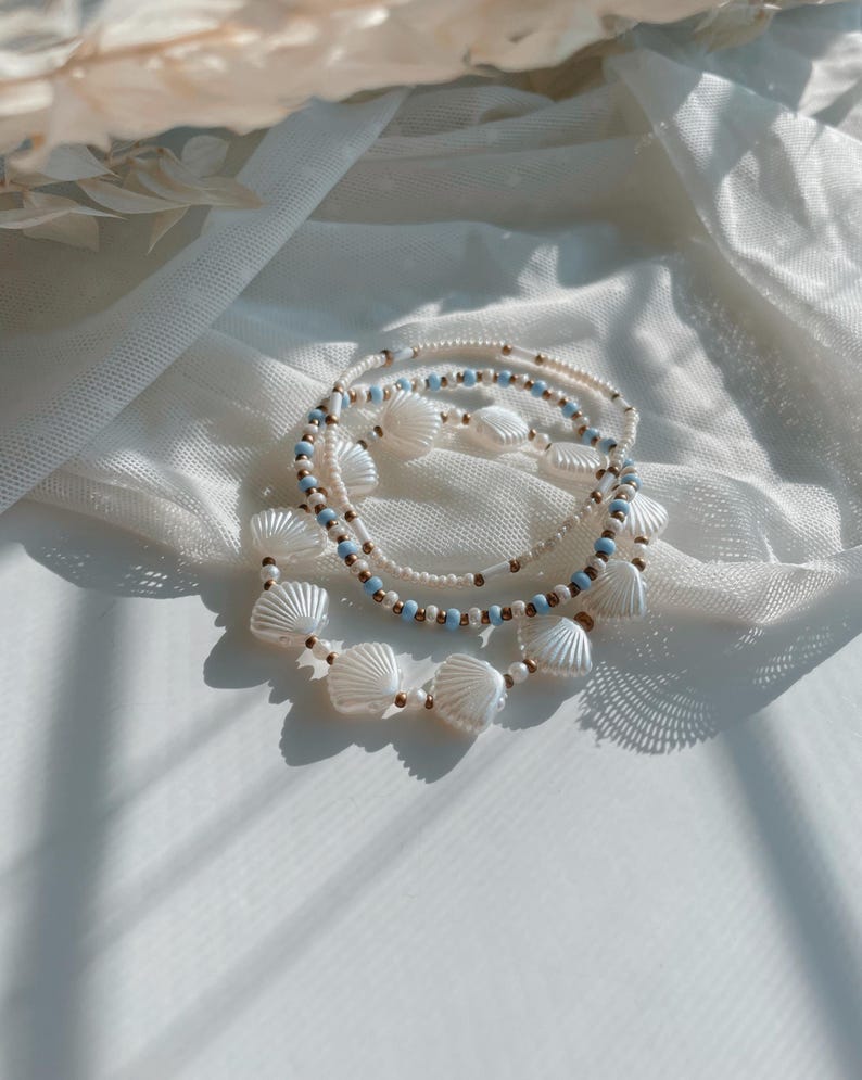 Peut inclure: Un ensemble de trois bracelets de perles compos&eacute; de perles blanches en forme de coquillage, de petites perles rondes bleu clair et de minuscules intercalaires dor&eacute;s m&eacute;talliques. Les bracelets sont dispos&eacute;s en cercle, r&eacute;v&eacute;lant des textures vari&eacute;es.