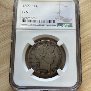 May include: A silver coin from 1899, graded G6, encased in a clear protective holder. The coin features a profile of a woman and the text "IN GOD WE TRUST". The holder includes the text "NUMISMATIC GUARANTY COMPANY".