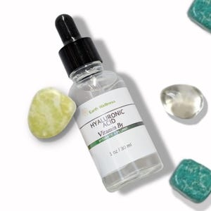 May include: A clear glass bottle of Earth Wellness Hyaluronic Acid Vitamin B5 serum with a black dropper top. The label reads "HYDRATE + PLUMP" and "1 oz / 30 ml". The bottle is surrounded by decorative stones in shades of green and clear.