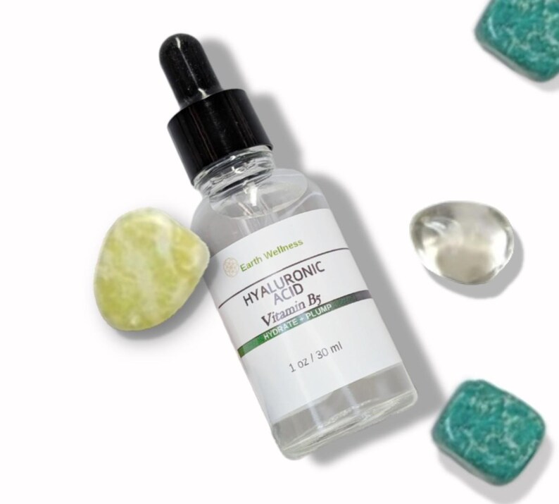 May include: A clear glass bottle of Earth Wellness Hyaluronic Acid Vitamin B5 serum with a black dropper top. The label reads "HYDRATE + PLUMP" and "1 oz / 30 ml". The bottle is surrounded by decorative stones in shades of green and clear.