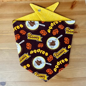 Op de afbeelding: Een bruine bandana met een gele rand. De bandana heeft een herhalend patroon van het San Diego Padres-logo, de mascotte van het team en de woorden "San Diego Padres".