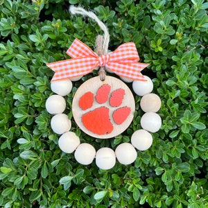 May include: A wooden bead wreath with a round orange paw print ornament in the center. The wreath is decorated with a white and orange gingham bow.