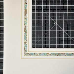 May include: A white mat board featuring a decorative border with a pattern of light blue and teal floral shapes on a tan background, accented by thin gold lines, positioned over a black cutting mat with a white grid and a 90-degree angle mark.