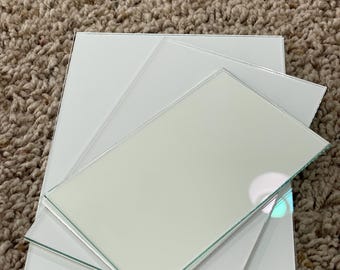 May include: A stack of square, white glass tiles with beveled edges. The tiles are arranged in a layered fashion, with the top tile slightly offset, revealing the reflective surface.