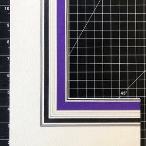 Puede incluir: Primer plano de una esquina de una estera de corte de cuadrícula negra y blanca con una cartulina de montaje en capas de color púrpura, negro, blanco y gris. La cartulina de montaje está cortada en un ángulo de 45 grados.
