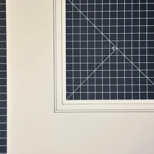 May include: A cream-colored mat board with a rectangular cutout, featuring a double-beveled edge. The mat is positioned over a dark gray cutting mat that displays a white grid pattern, including a 90-degree angle marking and thin white lines.
