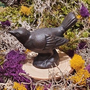 May include: A black cast iron bird figurine with detailed feathers, perched on a wooden base. The bird is surrounded by dried moss and flowers.