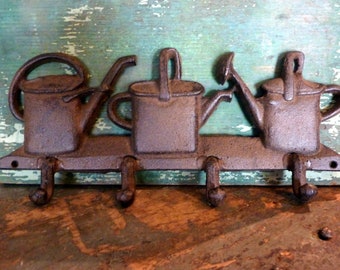 May include: A cast iron wall mounted key holder with three hooks shaped like watering cans. The watering cans are rusty brown in color and have a rustic look.