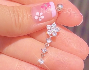 Peut inclure: Un piercing de corps argenté avec une chaîne pendante de petits strass clairs et un plus grand charme de fleur en strass.