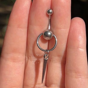 Op de afbeelding: Een zilverkleurige navelpiercing met een bungelende, puntige kegelvormige bedel. De ring heeft een kleine bal aan de bovenkant en een grotere bal in het midden, verbonden door een cirkelvormige ring. De kegelvormige bedel hangt aan de middelste ring.