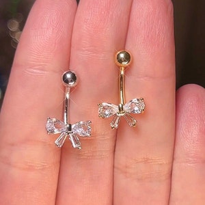 May include: Two belly button rings with a bow design. The bow is made of clear cubic zirconia stones. One ring is silver and the other is gold.