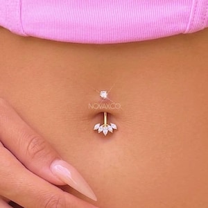 May include: A gold-colored belly button ring with a sparkling gem at the top and a cluster of small, white gems at the bottom. The ring is centered on a person's abdomen, with a pink top visible in the background.
