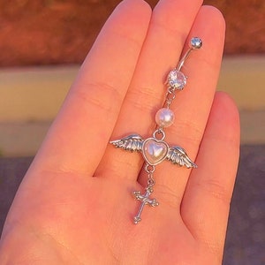 Puede incluir: Joyería corporal de plata con un dije de corazón con alas, una perla y un dije de cruz. La joyería se sostiene en una mano.