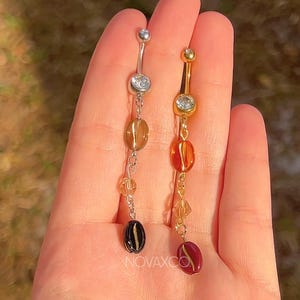 May include: Two belly button rings, one silver and one gold, each with a dangling chain of beads. The silver ring has a clear gem and a chain with amber, clear, and black beads. The gold ring has a clear gem and a chain with amber, clear, and red beads. The brand name "NOVAXCO" is visible.