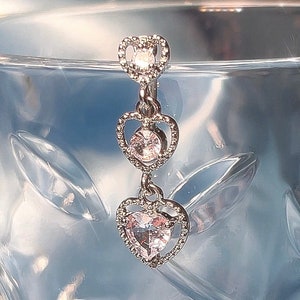 Pode incluir: Um piercing de umbigo prateado com três pingentes em forma de coração, cada um com um strass transparente no centro.