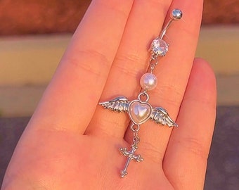 Peut inclure: Bijou de corps en argent avec un pendentif en forme de cœur avec des ailes, une perle et un pendentif en forme de croix. Le bijou est tenu dans une main.
