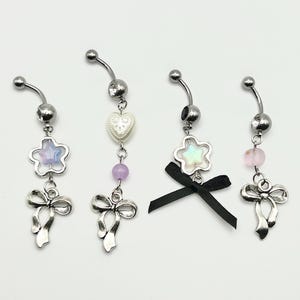 May include: Four silver belly button rings with different charms. The charms include a heart, a star, a bow, and a star with a bow. The charms are made of silver and have a white, pink, and purple color scheme.