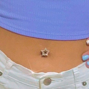 May include: A close-up of a person's midriff with a star-shaped belly button ring. The person is wearing a light blue crop top and white jeans.