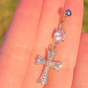 May include: A silver belly button ring with a clear crystal and a small pearl on a cross charm.