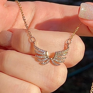 Op de afbeelding: Een goudkleurige ketting met een paar engelen vleugels, elke vleugel versierd met kleine, sprankelende kristallen. De ketting wordt door een persoon vastgehouden, waarbij de fijne ketting en het gedetailleerde ontwerp van de hanger worden benadrukt. De vleugels zijn ongeveer 2 cm breed.