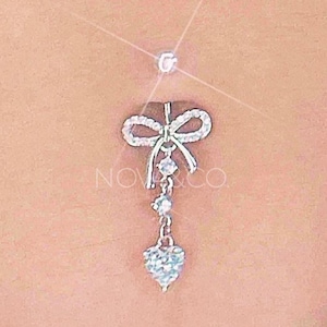 Puede incluir: Un piercing de ombligo plateado con un colgante de corazón y un lazo con brillantes transparentes.