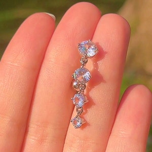 May include: A silver earring featuring five clear, round gemstones of different sizes. The earring hangs from a delicate chain, creating a cascading design. The background is a soft, blurred green.