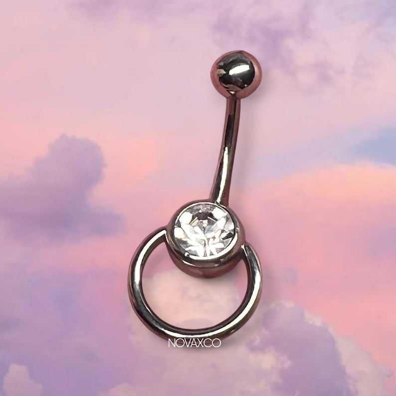 Peut inclure: Un piercing de nombril en forme de barre courbe de couleur or rose m&eacute;tallique. Le bijou pr&eacute;sente une petite perle sph&eacute;rique sur le dessus et un sertissage inf&eacute;rieur plus large contenant une pierre transparente ronde &agrave; facettes, entour&eacute;e d'un anneau.