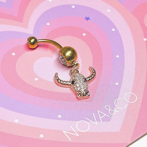 Puede incluir: Anillo para el ombligo dorado con un dije de toro. El dije de toro está incrustado con pequeños cristales transparentes. El anillo se muestra en una tarjeta en forma de corazón rosa y morado con el texto "NOVA & CO."