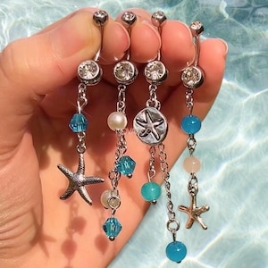 May include: Four silver belly button rings with dangling charms. Each ring has a clear gem at the top and a chain with beads and a charm. Charms include a starfish and a seashell. The beads are blue, white, and pink.