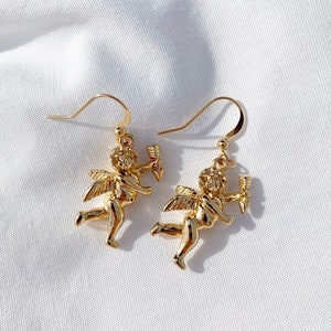 May include: A pair of gold-tone dangle earrings featuring a cherub design. Each cherub is holding an arrow and has wings. The earrings have a hook closure.