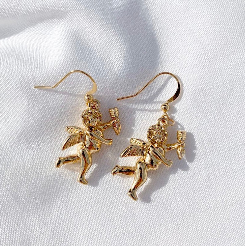 May include: A pair of gold-tone dangle earrings featuring a cherub design. Each cherub is holding an arrow and has wings. The earrings have a hook closure.