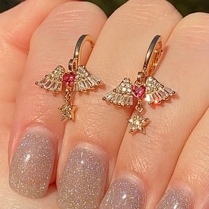 May include: Gold-coloured earrings with a celestial design. Each earring features a heart-shaped pink gem, surrounded by clear crystals and a dangling star charm. The earrings are held by a hand with glittery nail polish.