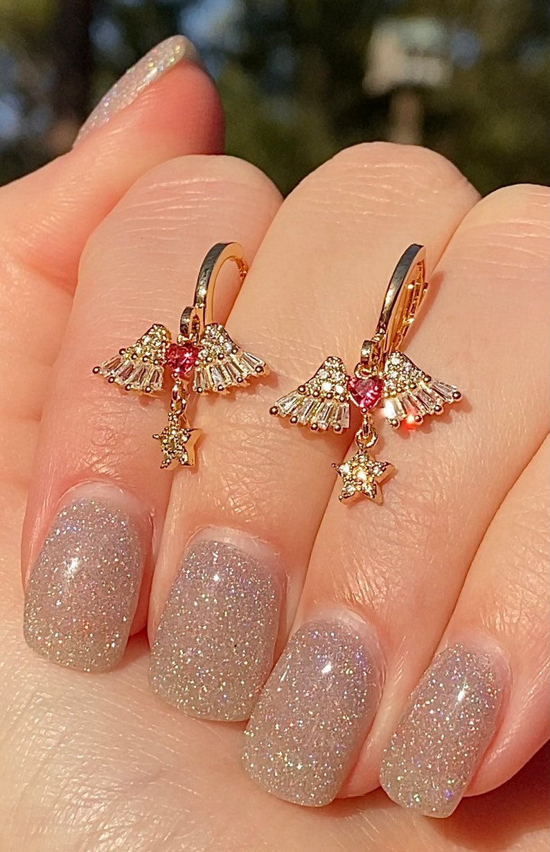 May include: Gold-coloured earrings with a celestial design. Each earring features a heart-shaped pink gem, surrounded by clear crystals and a dangling star charm. The earrings are held by a hand with glittery nail polish.