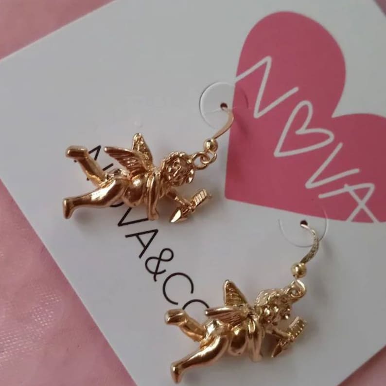 May include: Gold-tone earrings featuring a cherub design with a heart-shaped arrow. The earrings are on a white card with the text "NIVA" and "NA&CO" printed on it.