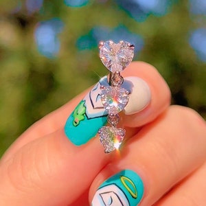 May include: A silver belly button ring with three heart-shaped cubic zirconia stones. The ring is held by a hand with a light blue and white manicure. The background is a blurred green.