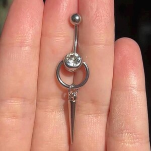 Peut inclure: Un piercing de nombril argent&eacute; compos&eacute; d'une barre courbe avec une petite boule sup&eacute;rieure et une grosse pierre ronde transparente &agrave; la base. Un anneau m&eacute;tallique est fix&eacute; &agrave; la pierre, suspendant une longue breloque en forme de c&ocirc;ne pointu.